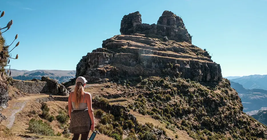 Mulher olhando para a fortaleza de Waqrapukara em Cusco, Peru, com um céu azul claro e muita vegetação ao redor da fortaleza