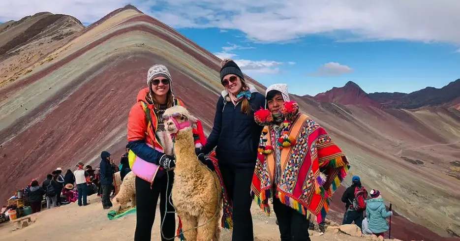 Turistas posando em Vinicunca com uma lhama e uma criança, todos usando chapéus, um céu azul com algumas nuvens e montanhas ao fundo.